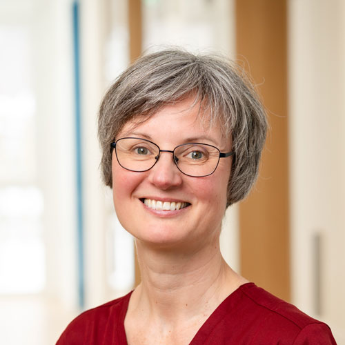 Porträt von Katrin Beckmann, Fachärztin für Kinder- und Jugendmedizin mit Schwerpunkt Kinderkardiologie in Minden. Sie hat kurze grau melierte Haare, trägt eine schmale Brille und ein rotes Oberteil. Das Foto zeigt sie vor einem hellen Praxis-Hintergrund.