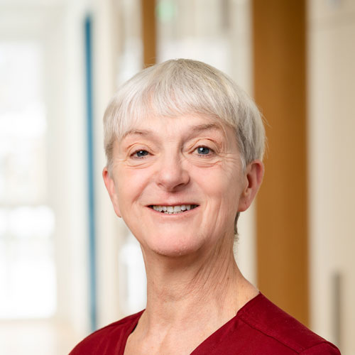 Porträt von Ute Becker, Fachärztin für Kinder- und Jugendmedizin mit Schwerpunkt Neonatologie. Sie hat sehr kurze, hellgraue Haare und trägt ein rotes Oberteil. Das Foto zeigt sie vor einem hellen Praxis-Hintergrund.